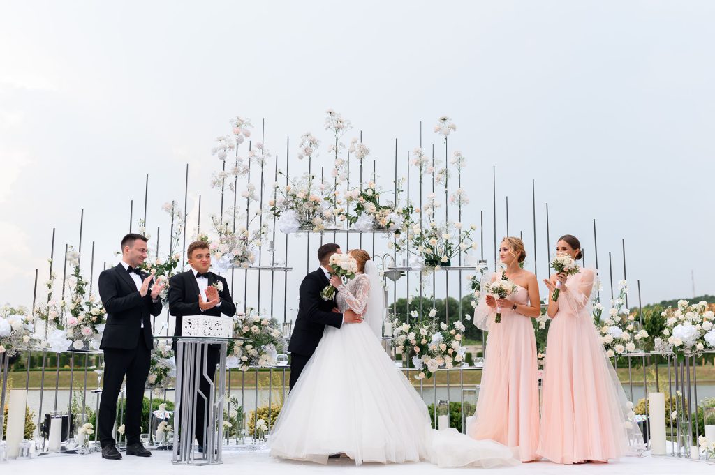 Front view of wedding ceremony outdoors, man in suits kissing woman in white puffy dress, while standing with bridesmaids and groomsmen near floral arch.